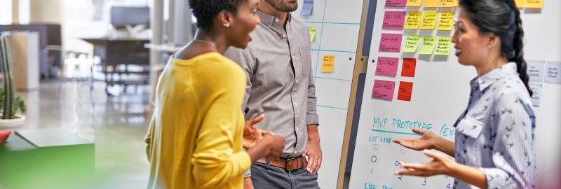 Three people have a conversation while standing near whiteboards with notes attached