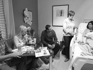 In a hospital room, a family is visiting a woman that is in bed, they are talking and drinking beverages.