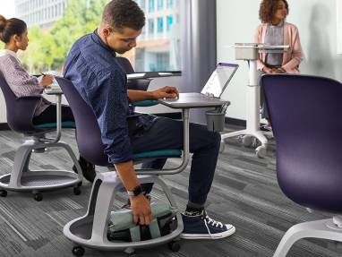 A student reaches for a backpack placed beneath the seat of a Node Tripod Base chair