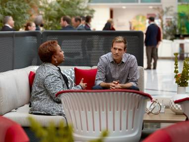 Two persons talking while seated on a grey sofa with red pillows