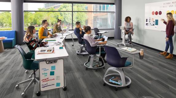 A group of students in a classroom sits in Node tripod base chairs and Node 5-star base chairs while a person at a whiteboard teaches