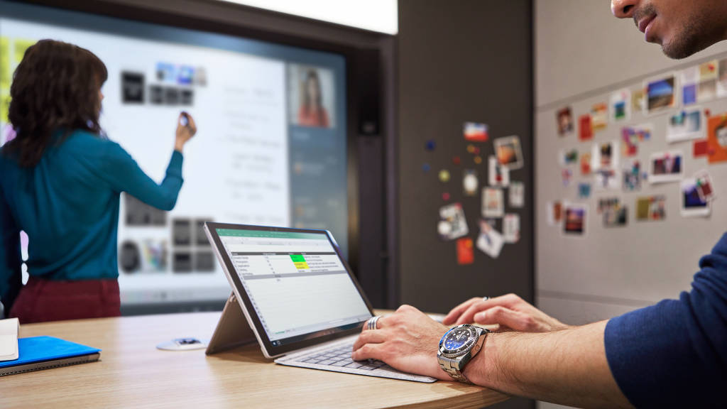 Hands are seen on a laptop's keywords while a person in the background presents information on a screen